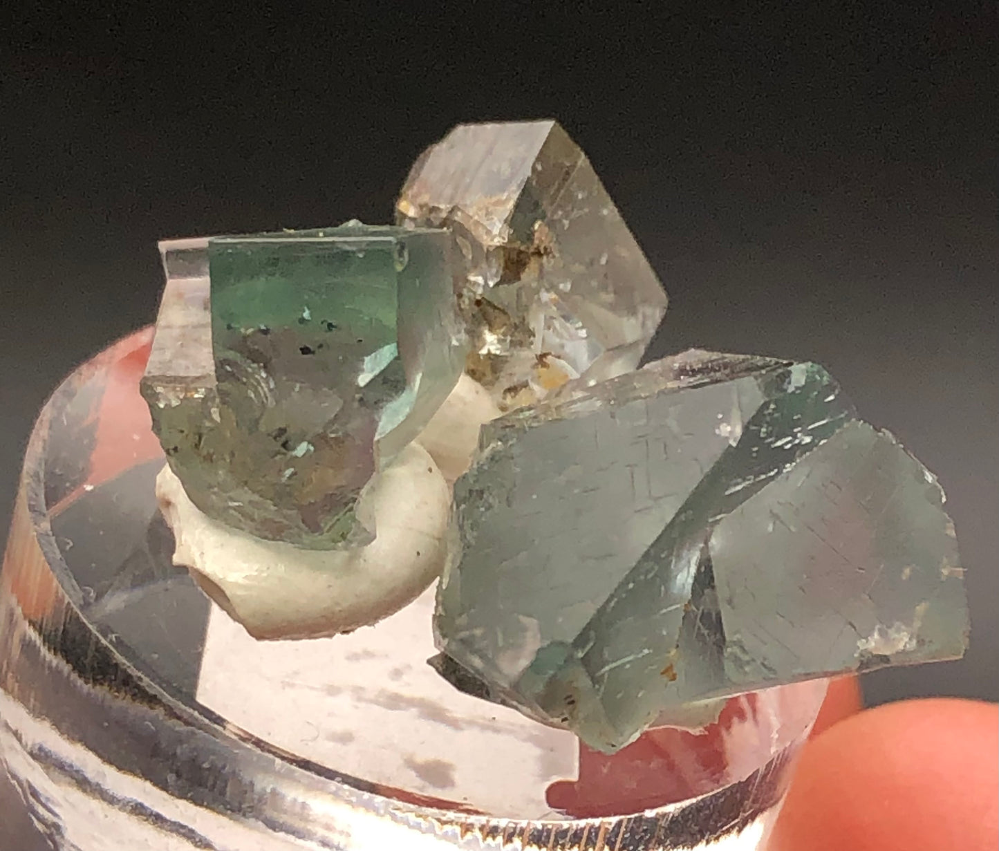 Close-up of three pale green, transparent fluorite formations from The Crystalary's "Rainbow's End Pocket" in Lady Annabella Mine, set against a dark background. The geometric crystals on a small stand highlight their clarity and distinct facets.