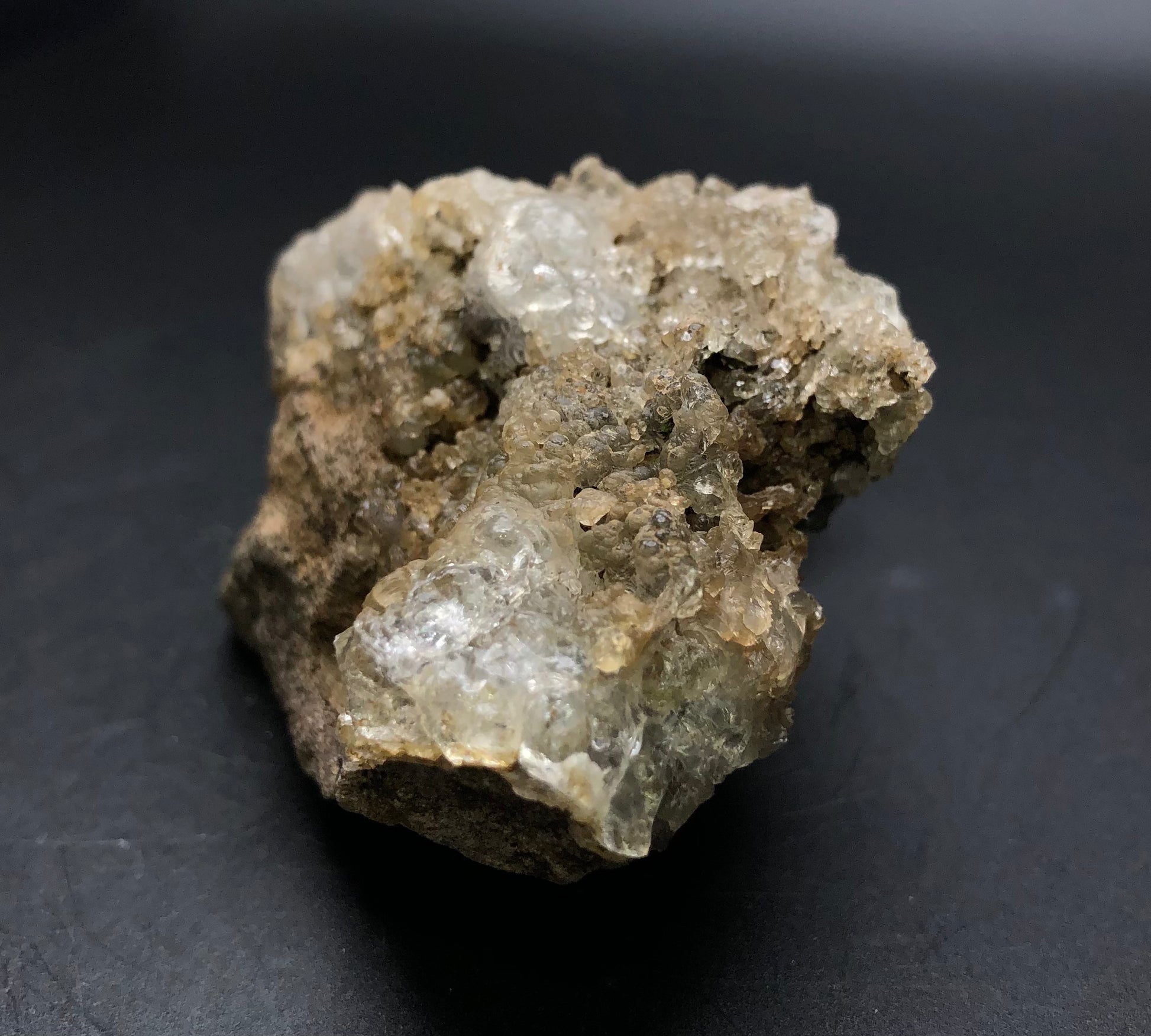 A rough, translucent stalactic hyalite opal from The Crystalary collection features a crystalline surface, resting on a dark, smooth background. Originating from La Ventilla in the Villa de Reyes Municipality of San Luis Potosí, Mexico, this mineral showcases beige and gray tones with sparkling facets that reflect light and exhibit a subtle green fluorescence.