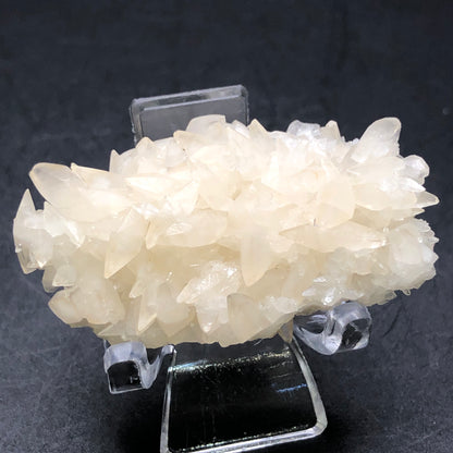 A display features a cluster of cream-colored calcite crystals resembling the renowned peach calcite from Southwest Mine, Hendricks Gulch, Bisbee, Arizona. The Crystalary presents these pointed translucent crystals on a clear stand against a dark background.