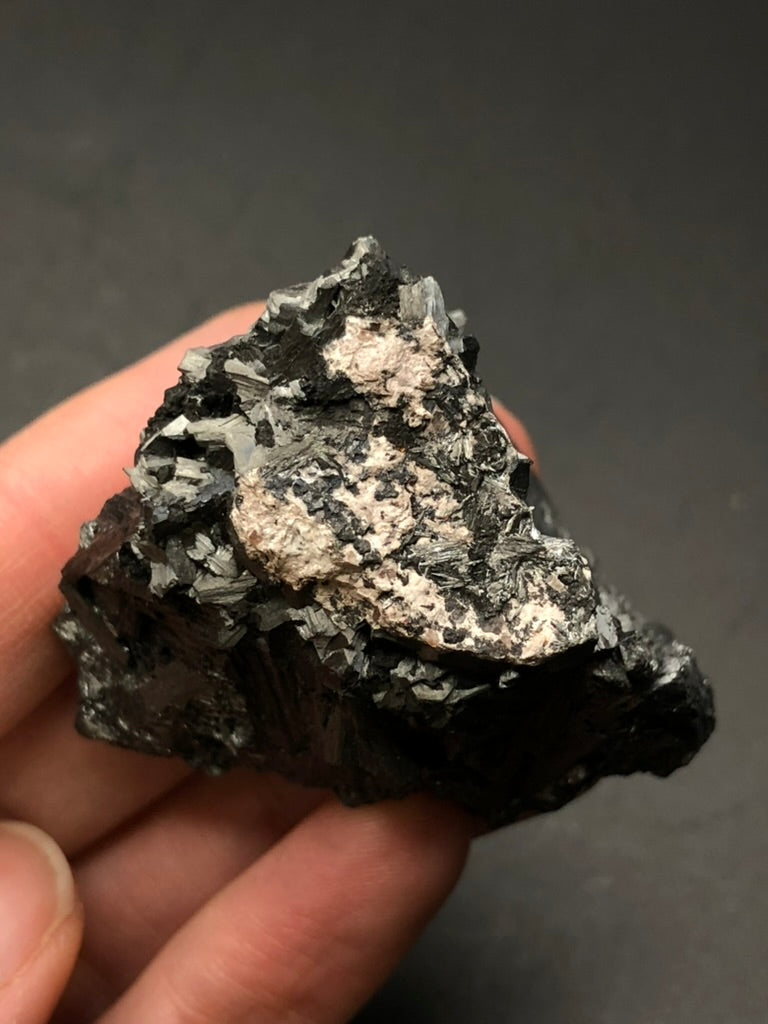 A hand holds a raw, dark AUCTION- Pyrolusite crystal from Elgersburg, Ilm District, Thuringia, Germany by The Crystalary. Its jagged edges and light pinkish patch are highlighted against a dark background.