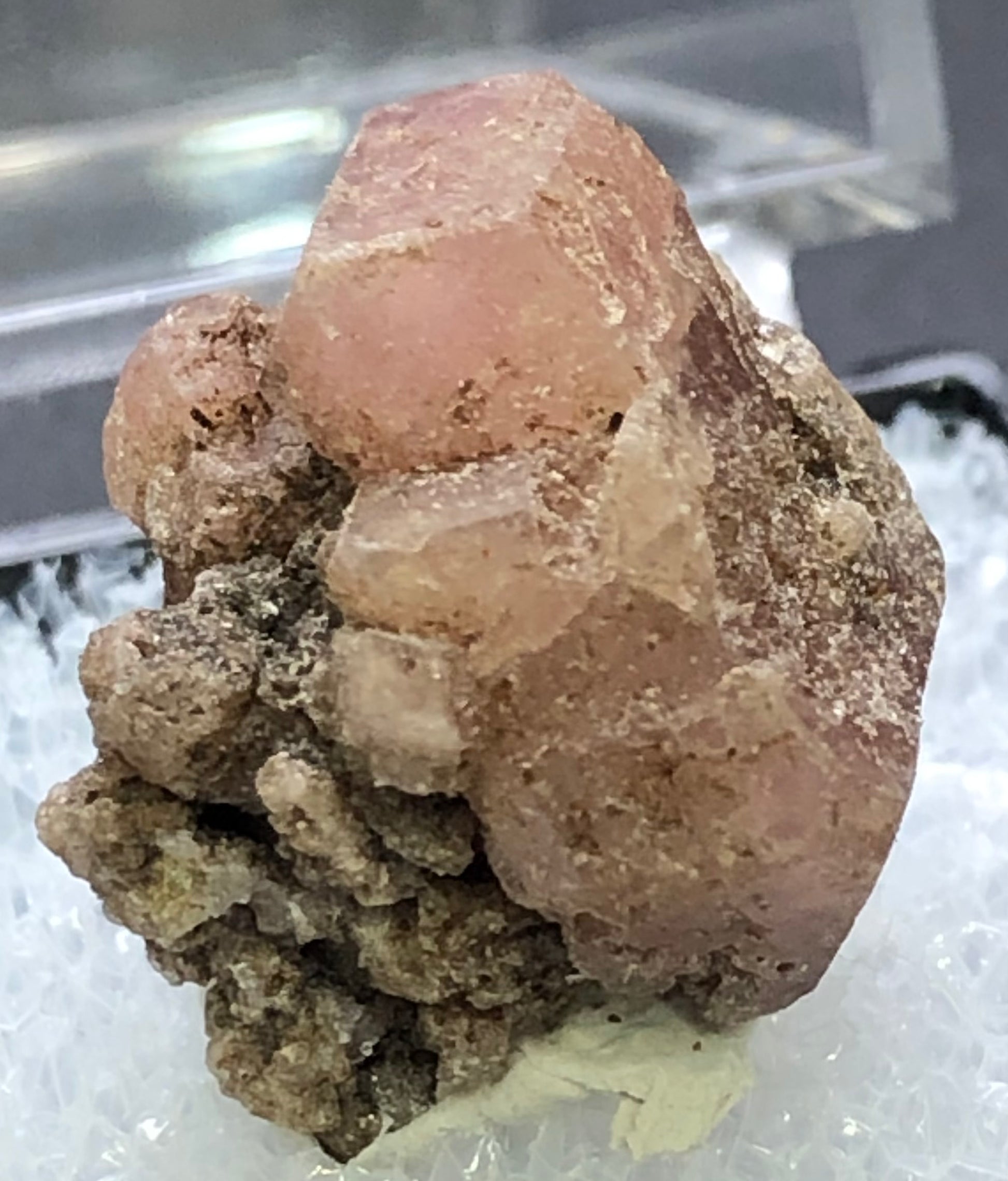 A close-up of The Crystalary's AUCTION- Garnet, Grossular from Morelos, Mexico, showcases a dusty rose pink cluster on a light background. The main crystal features a rough texture and irregular surfaces with smaller mineral formations surrounding it.