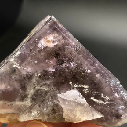 An up-close image features the AUCTION- Fluorite by The Crystalary, showcasing its rough, uneven surfaces and shimmering facets. This triangular formation against a dark background highlights the daylight color-changing hues that enhance its natural beauty.