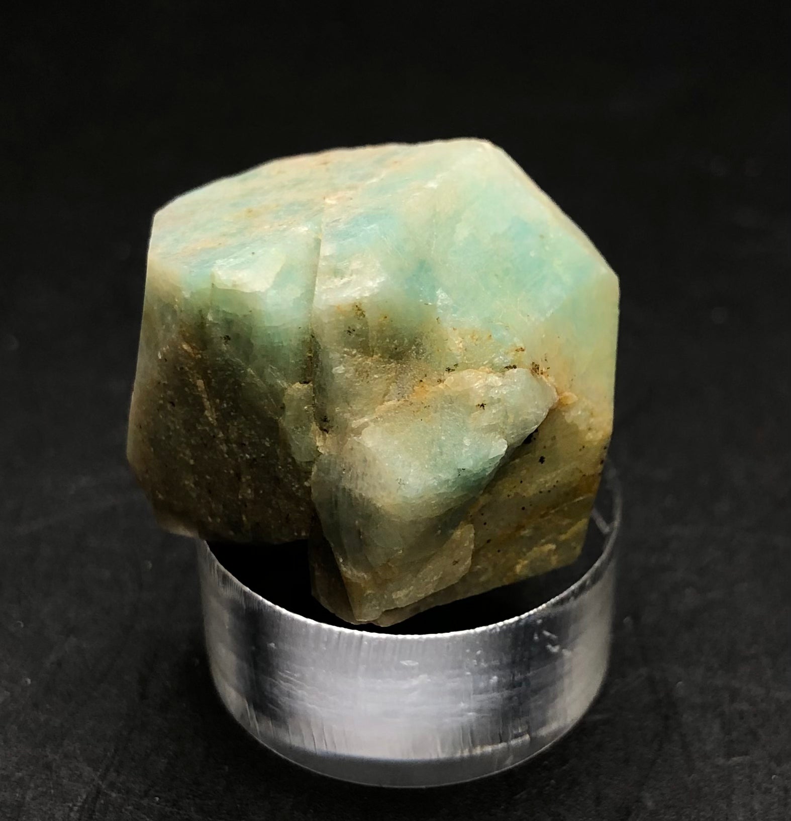 An Amazonite from Lake George, Park County, Colorado, USA, sourced by The Crystalary, features a rough and irregular shape with earthy green tones and a vitreous luster. It rests on a silver metallic cylindrical base against a black background.