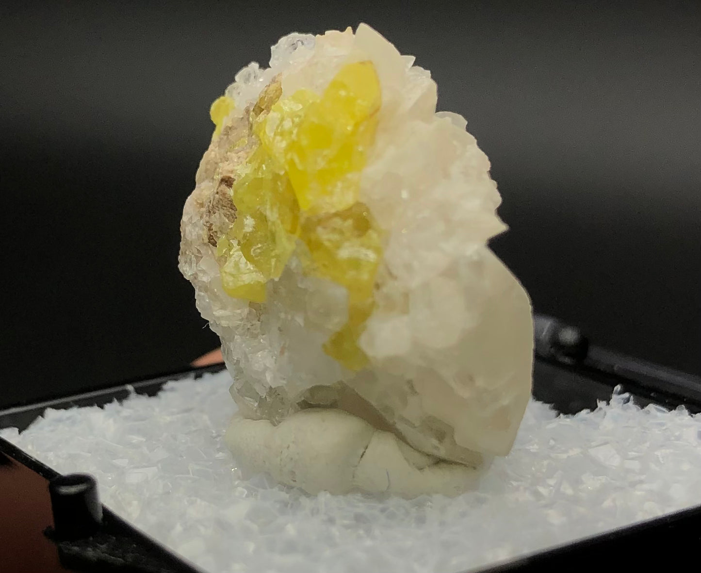 A detailed close-up of the AUCTION- Sulfur, Calcite mineral specimen from Maybee Quarry, offered by The Crystalary, features translucent white crystalline formations and vibrant yellow sulfur crystals set against a dark background. This striking mineral is elegantly displayed on a white base sprinkled with calcite crystals.