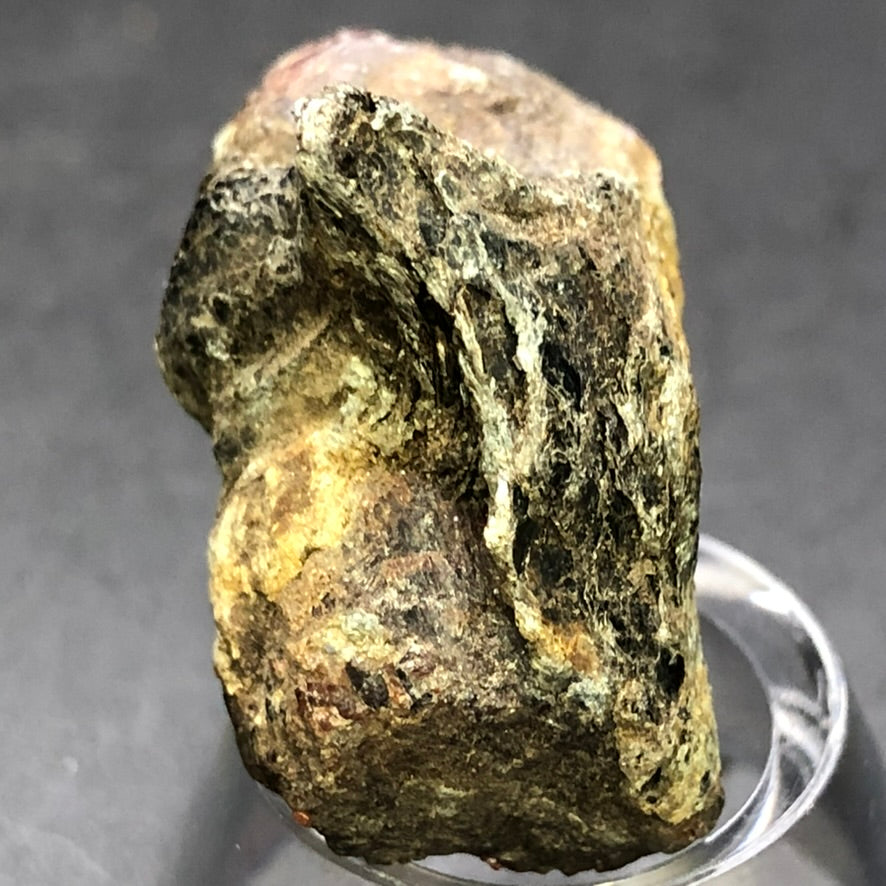 The Crystalary's AUCTION- Almadine Garnet from Markstay, Ontario, displays a rough, multicolored surface with a metallic sheen and shades of brown, green, and gray. It sits on a clear stand against a dark background, highlighting its diverse geological beauty.