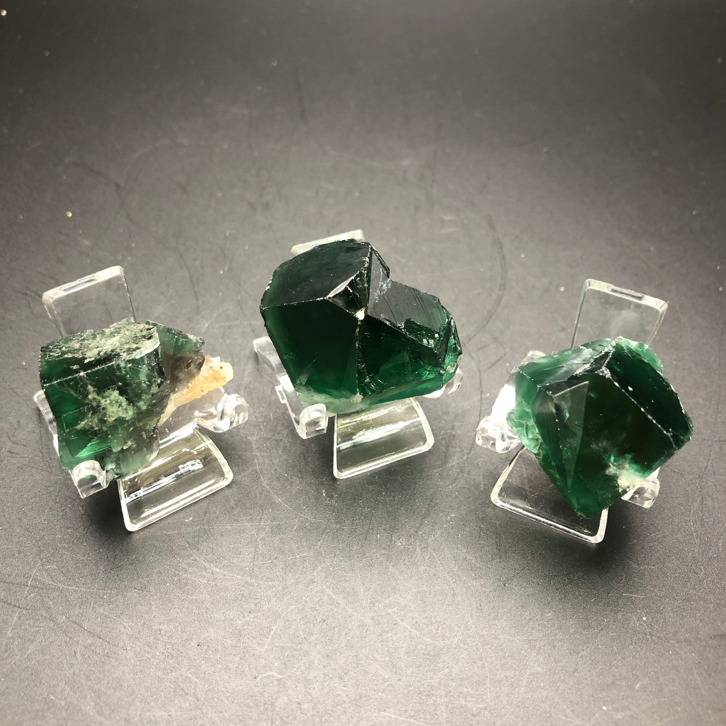 Displayed on clear stands against a dark background are a trio of deep green fluorite crystals from the Diana Maria Mine in Frosterley, Weardale Co., Durham, England. Offered by The Crystalary under the name "AUCTION- Fluorite," each crystal showcases unique, angular formations and varying shades of green that highlight their captivating beauty.