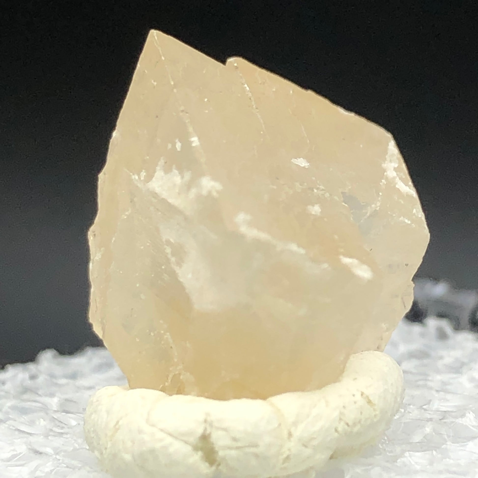 Close-up of a translucent, light honey Calcite crystal from Otero County, Colorado, displayed on a white base. The Crystalary offers this jagged-edged mineral with a textured surface against a dark backdrop.