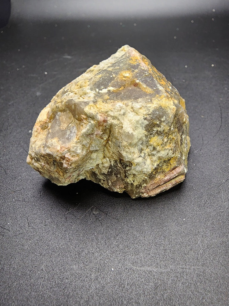 A rough, unpolished piece from The Crystalary’s Lazlo Dudas Collection features the Elbaite variety, Rubellite, hailing from Dobra Voda, Velke Mezirici in Moravia, Czech Republic. Resting on a dark surface, its mix of gray, yellow, and brown hues accentuate its irregular shape with varied textures and surfaces.