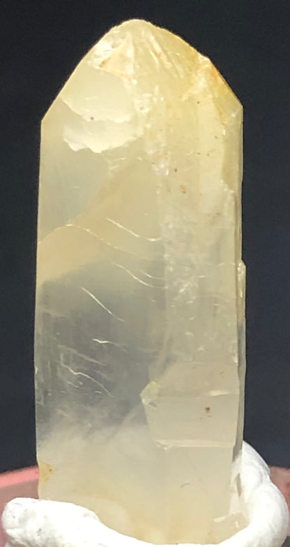 A close-up of a translucent, hexagonal Mango Quartz crystal from Cabiche, Colombia, shows smooth surfaces and visible striations. Displayed upright on a white base against a dark background, this piece is from The Crystalary's AUCTION- Quartz w/ Halloysite collection.