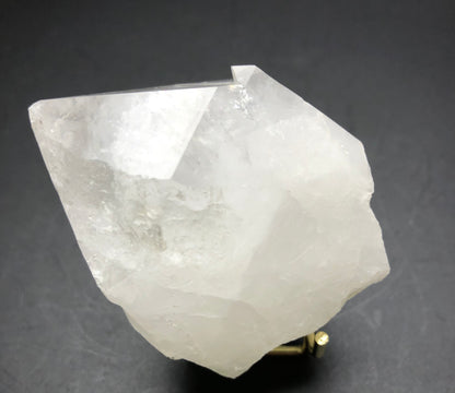 The Crystalary's AUCTION- Quartz- unk locality features a large, translucent quartz point with angular facets, set against a dark background to highlight its natural, rough texture and slight reflective sheen in impressive crystal dimensions.