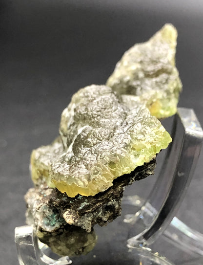 The Crystalary presents an AUCTION of a Smithsonite var. Cuprian from the 79 Mine in the Chilito, Hayden area of Banner Mining District, Gila County, Arizona. This greenish-yellow mineral with a rough, textured surface is elegantly showcased on a clear stand against a dark background. Its crystalline structure beautifully catches light on its uneven surfaces.