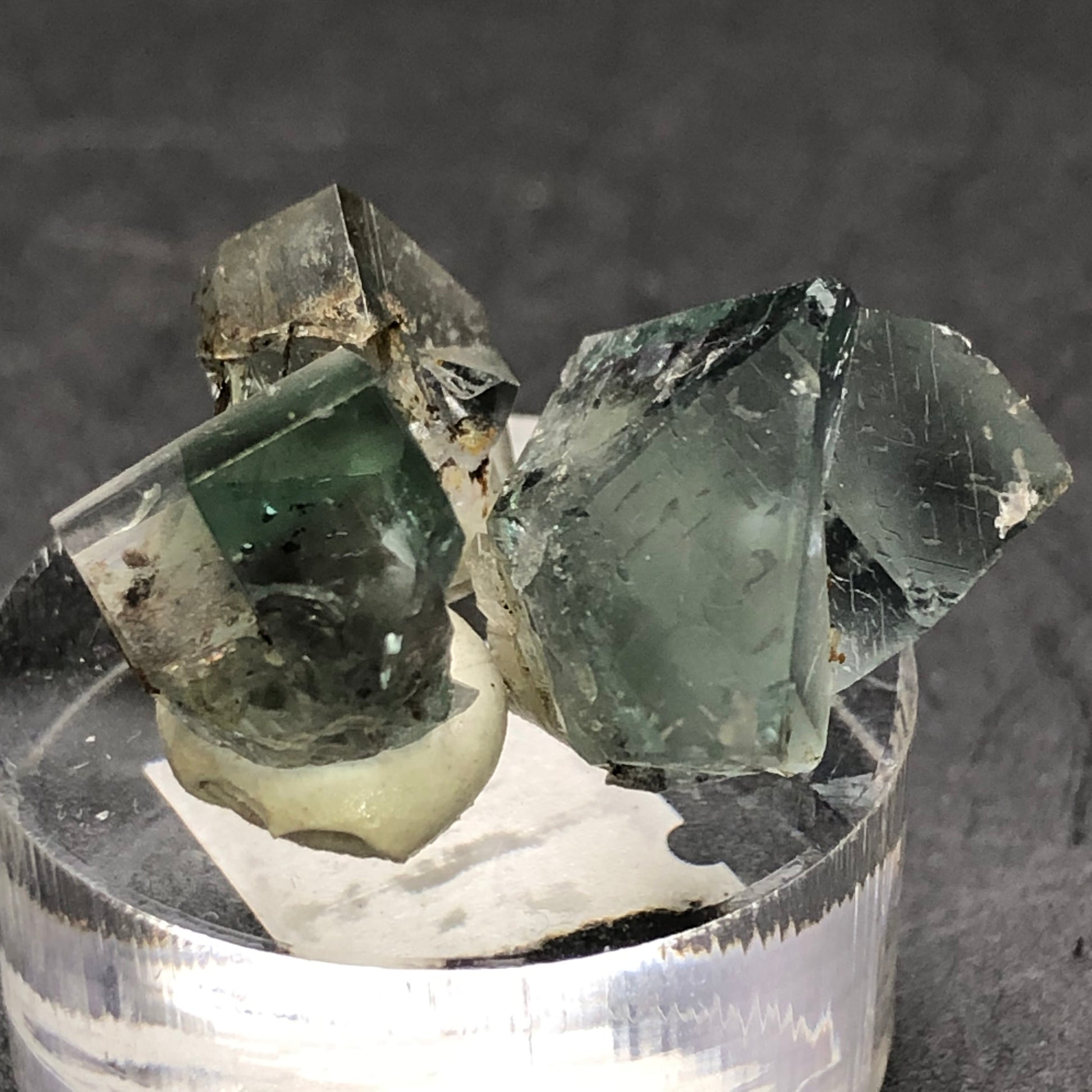 A close-up of three green fluorite crystals from The Crystalary's "AUCTION- Fluorite- Rainbow's End Pocket" showcases the Lady Annabella mine gems with smooth, angular surfaces on a clear cylindrical base against a dark background, highlighting their color and texture variations.