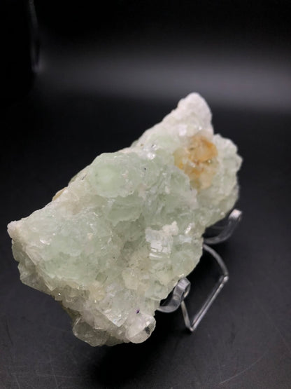 A close-up of a pale green AUCTION- Fluorite from Xianghualing Mine, featured by The Crystalary, is showcased on a clear stand against a dark background. The fluorite possesses a rough and uneven surface with translucent and white areas.