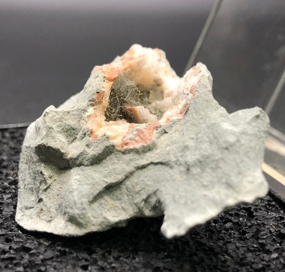 The Crystalary's AUCTION-Millerite product features a mineral rock with a greenish-grey exterior. Its hollow center is adorned with fibrous, pale green millerite crystals and white crystalline structures, resembling a druzy quartz geode, all beautifully presented on a textured black surface.