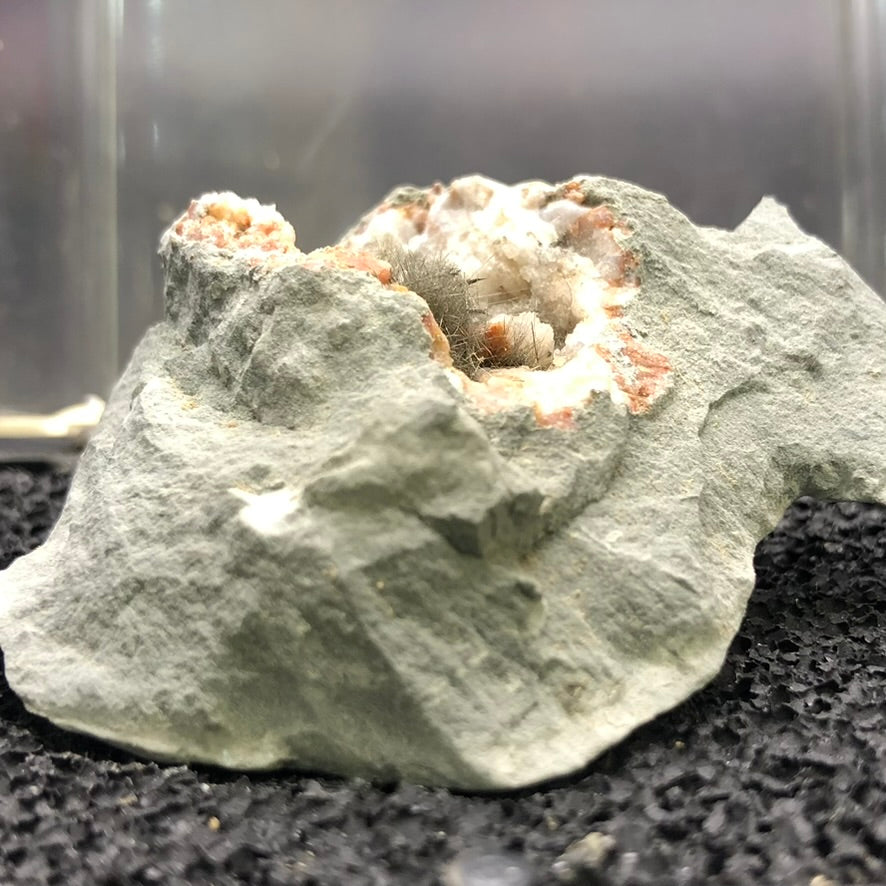 A close-up view of the "AUCTION- Millerite- locality unknown" by The Crystalary showcases a mineral specimen with a rough gray exterior, featuring a cluster of sparkling, silvery crystals that resemble Millerite's hair-like formations. These are surrounded by brownish-red and white deposits, and the piece is set against a textured black surface, echoing the captivating allure of a druzy quartz geode.