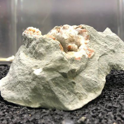 A close-up view of the "AUCTION- Millerite- locality unknown" by The Crystalary showcases a mineral specimen with a rough gray exterior, featuring a cluster of sparkling, silvery crystals that resemble Millerite's hair-like formations. These are surrounded by brownish-red and white deposits, and the piece is set against a textured black surface, echoing the captivating allure of a druzy quartz geode.