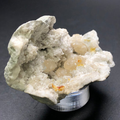 A close-up of "AUCTION- Geode with Calcite- Worthington, Ohio, USA" by The Crystalary showcases rough white crystalline formations adorned with small, translucent honey calcite crystals. The specimen is elegantly displayed on a small clear stand against a dark background, echoing the stunning discoveries from Worthington, Ohio.