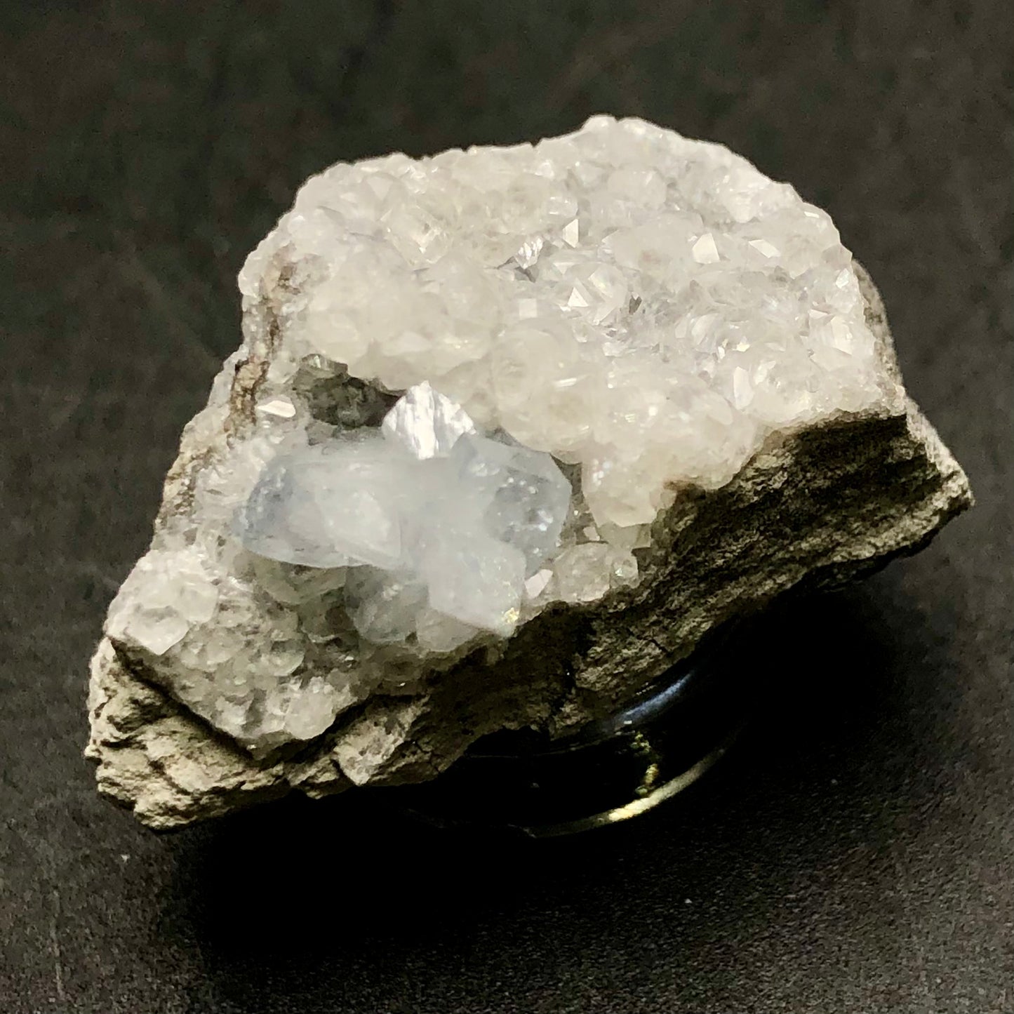 A rough, gray mineral rock from The Crystalary's collection at Meckley's Quarry in Mandata, Northumberland County, Pennsylvania features clusters of small, sparkling celestine crystals on its surface, set against a dark background. These crystals are primarily transparent with a subtle blue tint in certain regions.