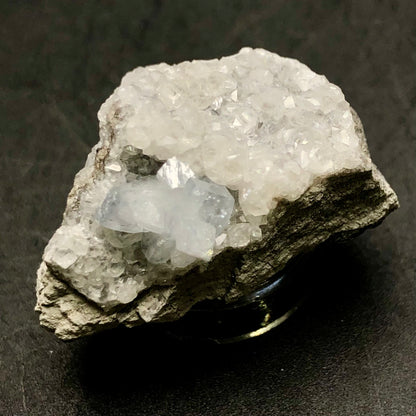 A rough, gray mineral rock from The Crystalary's collection at Meckley's Quarry in Mandata, Northumberland County, Pennsylvania features clusters of small, sparkling celestine crystals on its surface, set against a dark background. These crystals are primarily transparent with a subtle blue tint in certain regions.