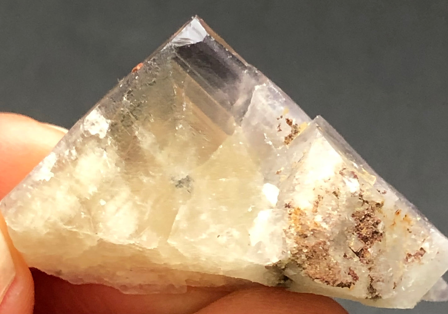 A hand holds a translucent Angular Fluorite Crystal from The Crystalary, featuring a pale yellow hue with darker inclusions and a golden interior. Its rough texture contrasts against a plain gray background. Origin: Cromwell Pocket, Greenlaws Mine, Daddry Shield, Stanhope, County Durham, UK.