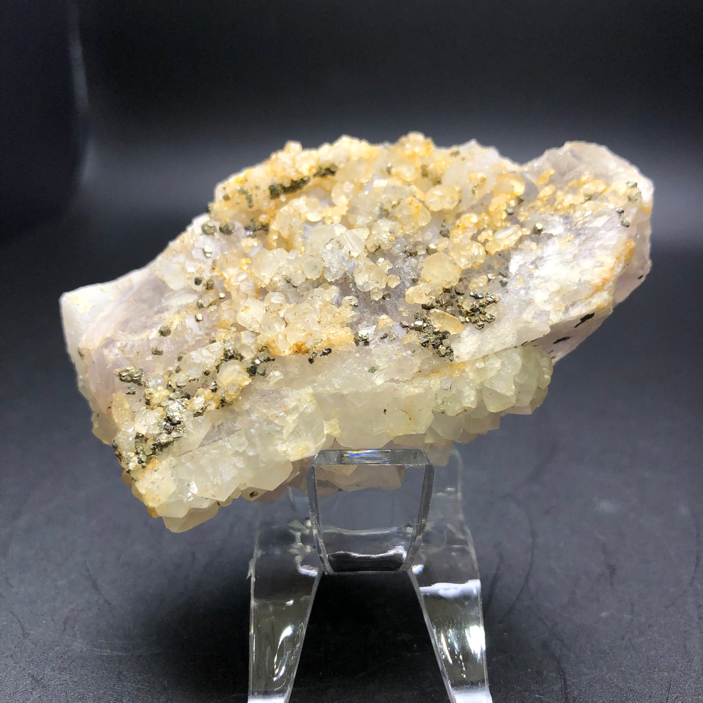 Introducing the unique offering from The Crystalary: the AUCTION-Fluorite, sourced from an unknown Chinese locality. This exquisite specimen showcases a base of translucent druzy quartz adorned with gleaming clusters of small, sparkling golden-yellow pyrite crystals. It is elegantly presented on a clear stand set against a dark backdrop to enhance its radiant beauty.