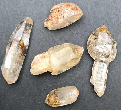 Five AUCTION-Petroleum Quartz crystals from The Crystalary, featuring varying sizes and shapes including a scepter, are showcased on a dark surface. These translucent Afghani crystals, some UV reactive with brownish inclusions, softly reflect light.