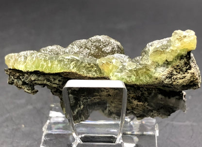 The Crystalary presents an intricate Smithsonite var. Cuprian specimen from the 79 Mine in the Chilito, Hayden area, Banner Mining District, Gila County, Arizona. This exquisite piece features green, translucent crystals resting on a dark base and is beautifully displayed on a clear stand against a dark background.