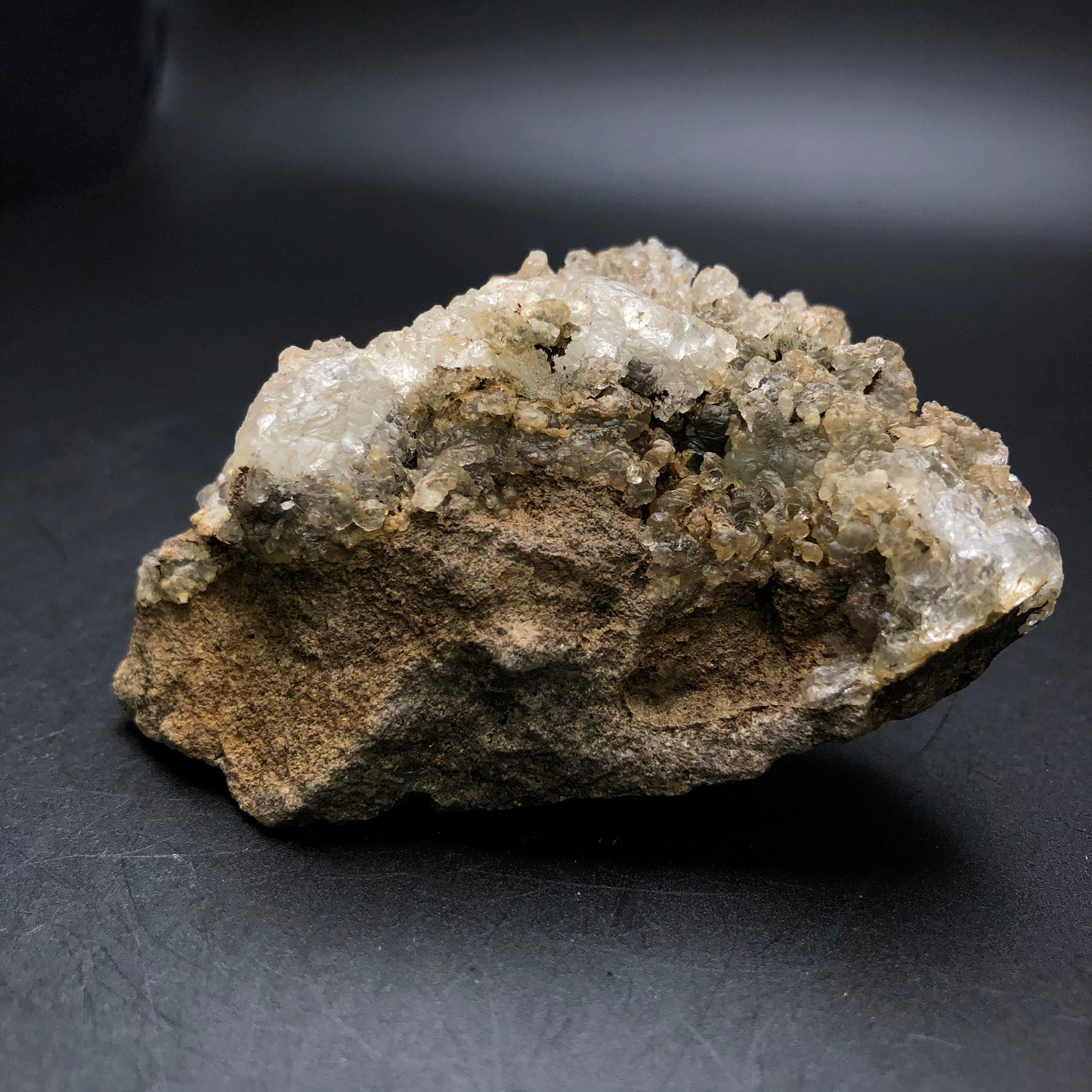 A rough brown and white mineral rock from The Crystalary, labeled as AUCTION- Hyalite- La Ventilla, Villa de Reyes Municipality, San Luis Potosí, Mexico, is set against a dark background. Its visible crystal formations glimmer gently under the light, revealing a subtle hint of green fluorescence reminiscent of stalactic hyalite opal from San Luis Potosí.