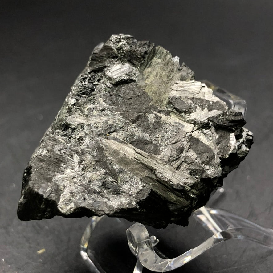 A close-up image of a dark, jagged specimen from The Crystalary brand features metallic and shiny streaks, likely pyrolusite. Displayed on a clear stand against a black background, it showcases its textured and layered surface with radiating crystals reminiscent of those found in Elgersburg. This piece is from the AUCTION collection: Pyrolusite - Elgersburg, Ilm District, Thuringia, Germany.