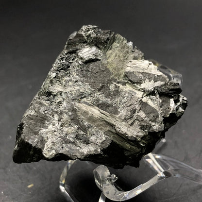 A close-up image of a dark, jagged specimen from The Crystalary brand features metallic and shiny streaks, likely pyrolusite. Displayed on a clear stand against a black background, it showcases its textured and layered surface with radiating crystals reminiscent of those found in Elgersburg. This piece is from the AUCTION collection: Pyrolusite - Elgersburg, Ilm District, Thuringia, Germany.