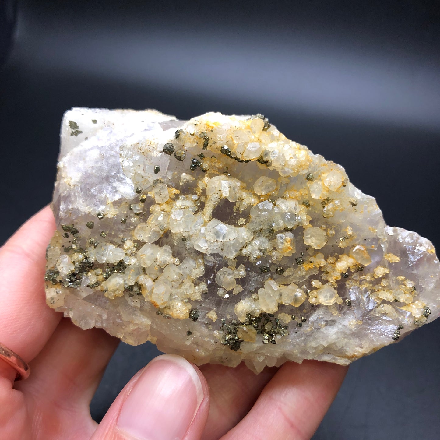 A hand holds a rough mineral rock, part of the AUCTION - Fluorite collection from The Crystalary, glistening with small druzy quartz crystals and metallic pyrite flecks against a dark background.