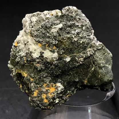 A close-up of a rough mineral with dark green, white, and yellowish-orange patches, evoking Hematite's earthy tones. The AUCTION- Pyrite and Hematite specimen from The Crystalary sits on a clear stand against a dark background.