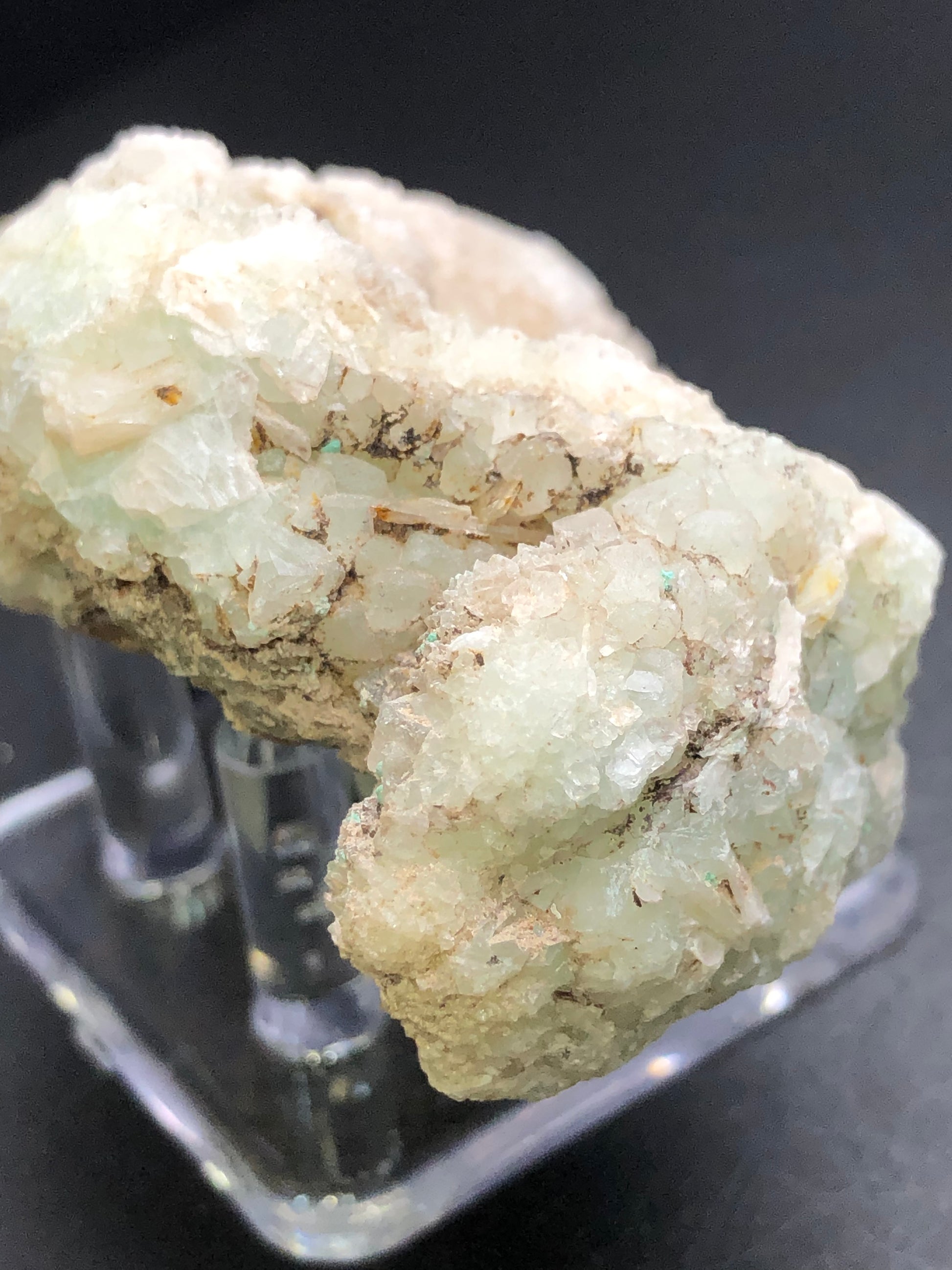 A detailed image showcases a rough, pale green mineral cluster from The Crystalary's AUCTION- Calcite, Quartz collection. Resembling druzy white quartz, the cluster with crystalline surfaces is set on a transparent stand against a dark background, highlighting its multiple small angular facets and textured surface.