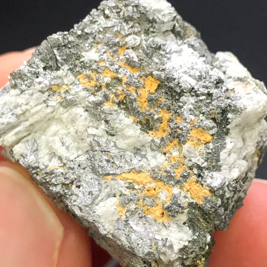 A hand holds "AUCTION- Pyrite, hematite- unk locality" by The Crystalary. The specimen's metallic texture and silver-gray with orange patches hint of oxidization, emphasized by a plain black background.