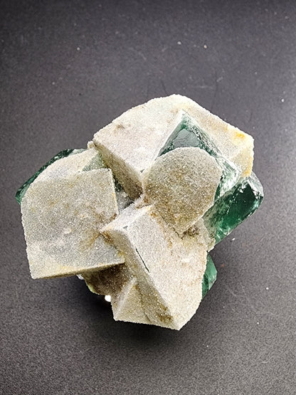 A cluster of translucent, emerald green Fluorite crystals from the Rogerley Mine in Frosterley, Weardale, Co. Durham, England, branded by The Crystalary as "Fluorite - Druzy Dreams Pocket," is partially covered by a beige, earthy mineral on a dark, textured background. The Fluorite crystals exhibit sharp edges and a smooth surface.