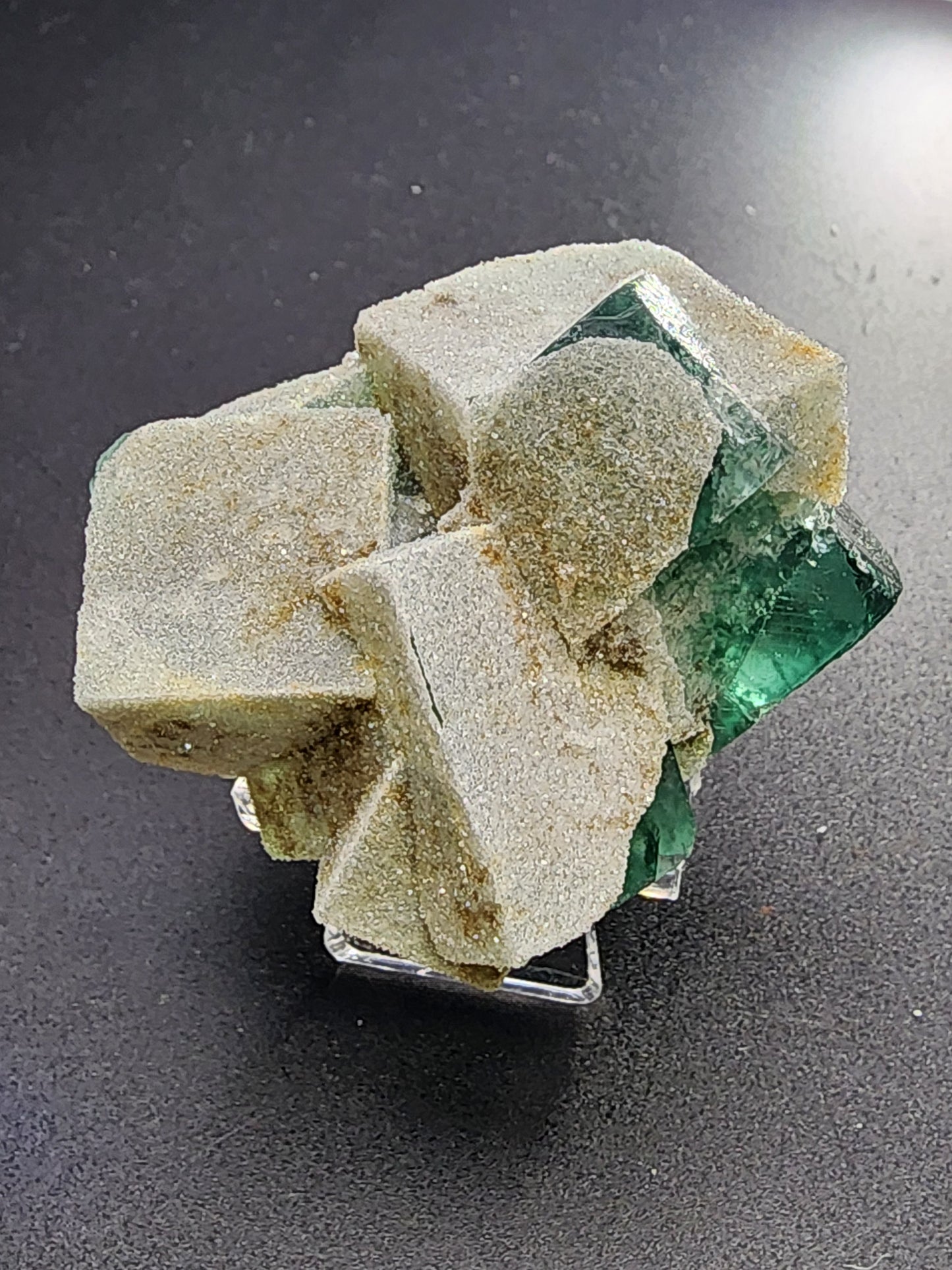 A cluster of cuboid emerald green fluorite crystals, partially covered with fine druzy quartz, from The Crystalary's Fluorite- Druzy Dreams Pocket at Rogerley Mine in Frosterley, Weardale, County Durham, England. The piece is displayed on a small, clear stand against a black background. The semi-translucent crystals exhibit visible edges and corners.