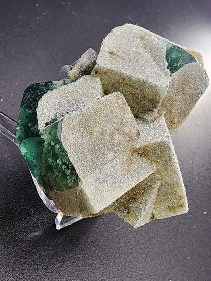 Close-up image of the Fluorite - Druzy Dreams Pocket from The Crystalary. This stunning emerald green fluorite crystal formation, sourced from the Rogerley Mine in Frosterley, Weardale, Co. Durham, England, features green crystals partially covered by powdery white Druzy quartz. The mix of sharp edges and rough surfaces creates a striking contrast against the dark, smooth background.