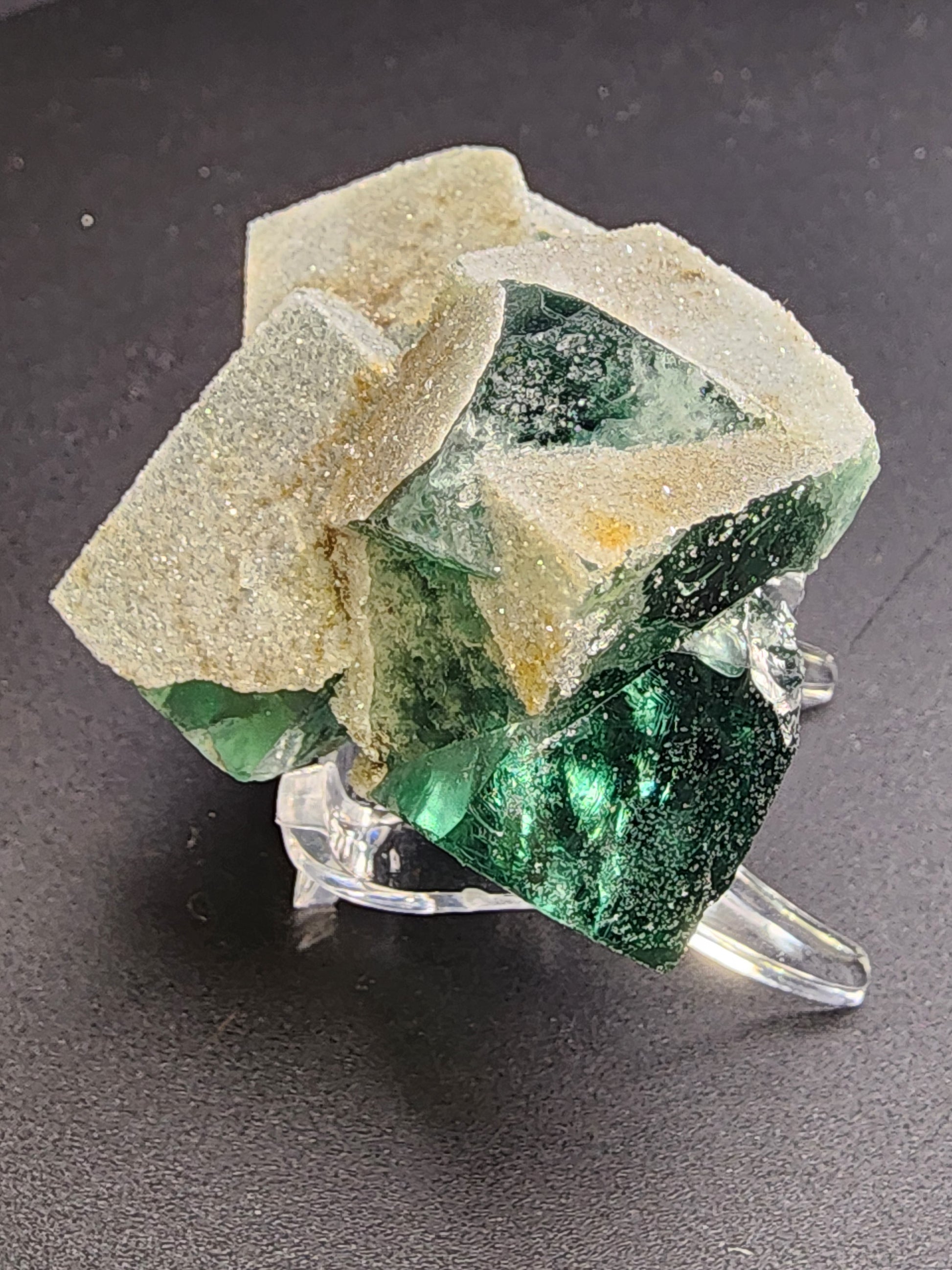 A close-up image showcasing "Fluorite- Druzy Dreams Pocket" from The Crystalary, featuring a stunning emerald green fluorite crystal cluster with cubic formations. The crystals have a sparkly surface reminiscent of druzy quartz and are mounted on a clear display stand set against a dark background. Originating from Rogerley Mine, Frosterley, Weardale, Co. Durham, England.