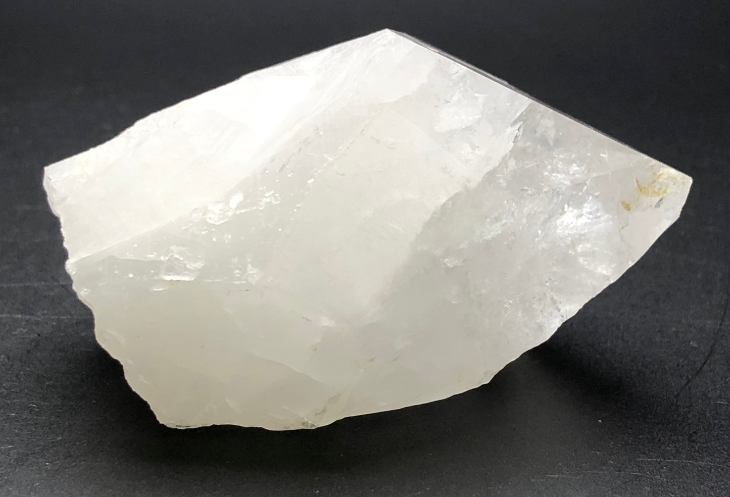 A rough, translucent quartz point from The Crystalary's "AUCTION- Quartz- unk locality" rests on a dark surface. Its irregular shape and natural facets exude a matte texture, highlighting subtle transparency.