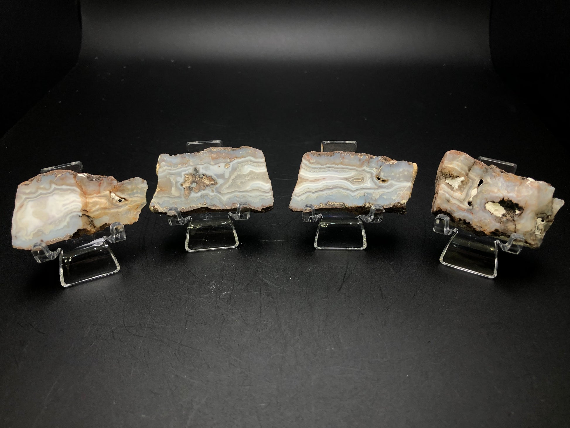 Four slabs of AUCTION- Agate from Fourth of July Peak, Hassayampa in Maricopa County, Arizona are elegantly displayed on small clear stands against a dark background. These agate pieces, offered by The Crystalary, feature intricate patterns and layers in shades of gray, white, and beige that showcase natural textures reminiscent of the Fourth of July Peak landscape.