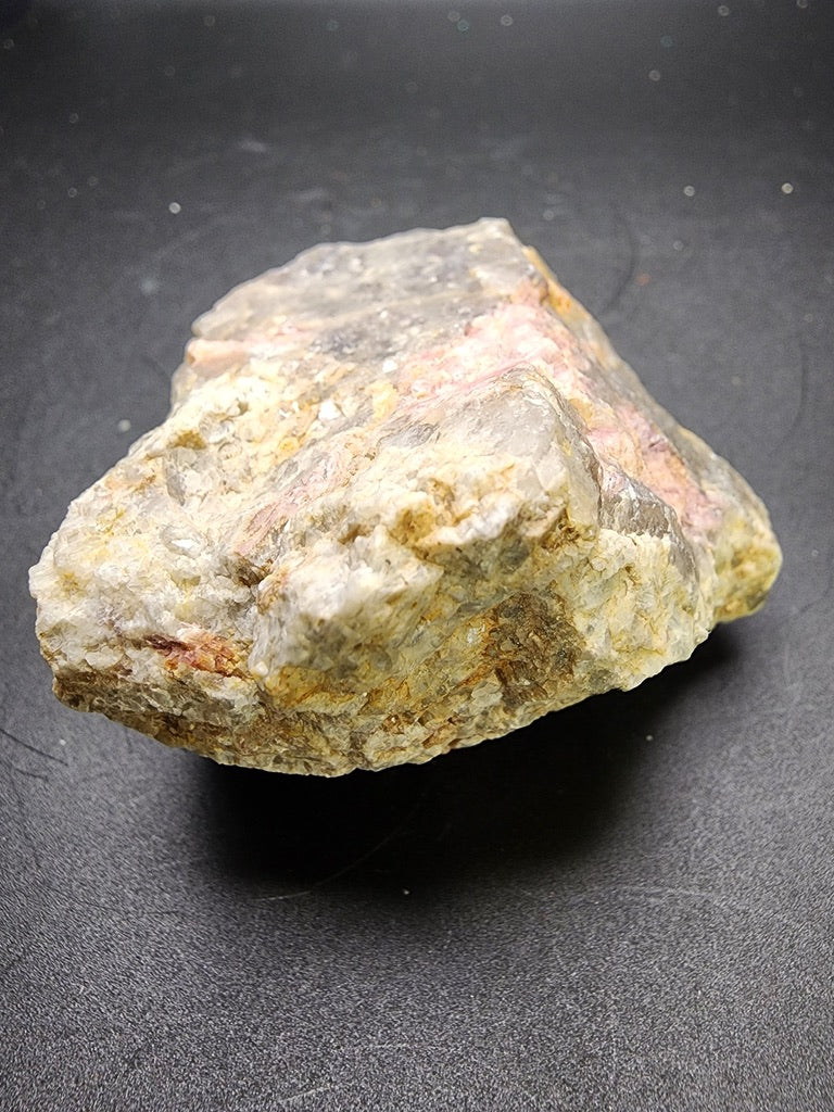 A raw, unrefined specimen from The Crystalary's AUCTION- Elbaite var. Rubellite features a blend of gray, white, and light pink tones, reminiscent of rubellite, set against a dark background surface.