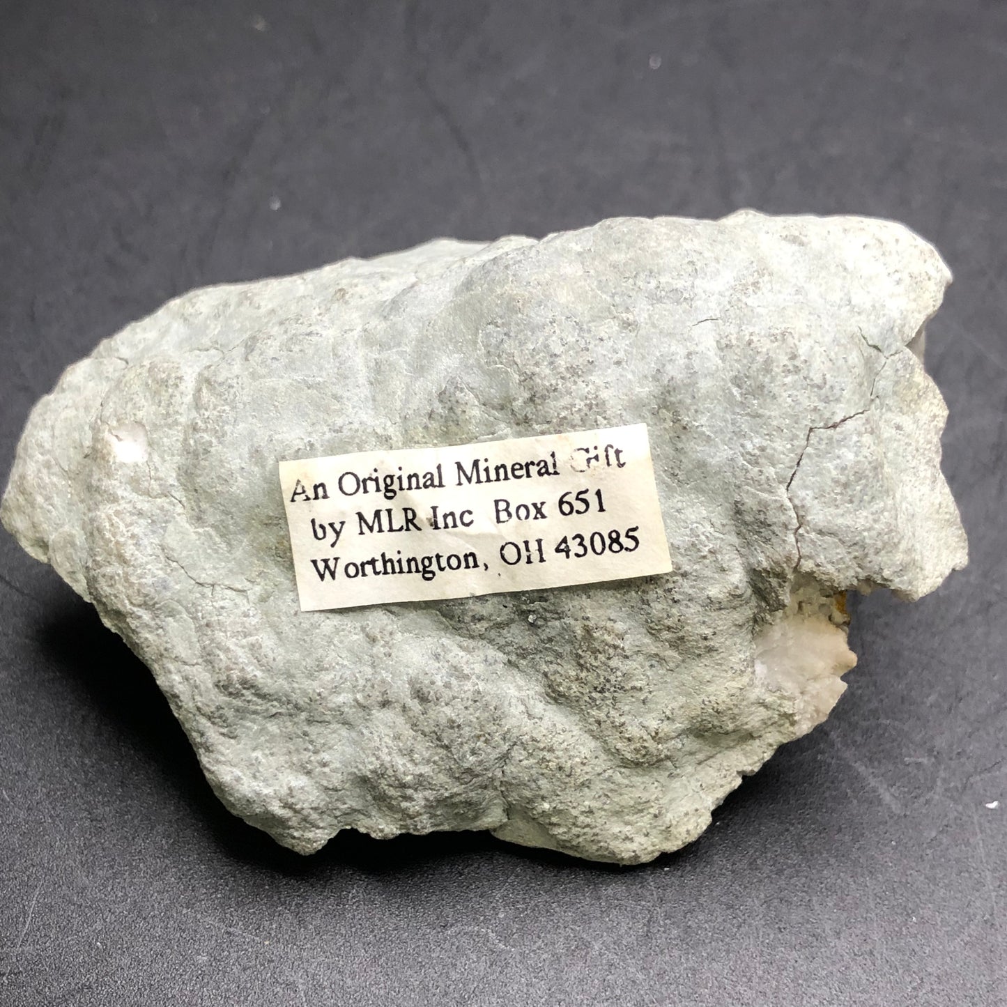 A grayish geode featuring calcite, branded with a label that reads, "The Crystalary," against a dark background. The packaging indicates it's an original mineral gift from Worthington, Ohio, USA.