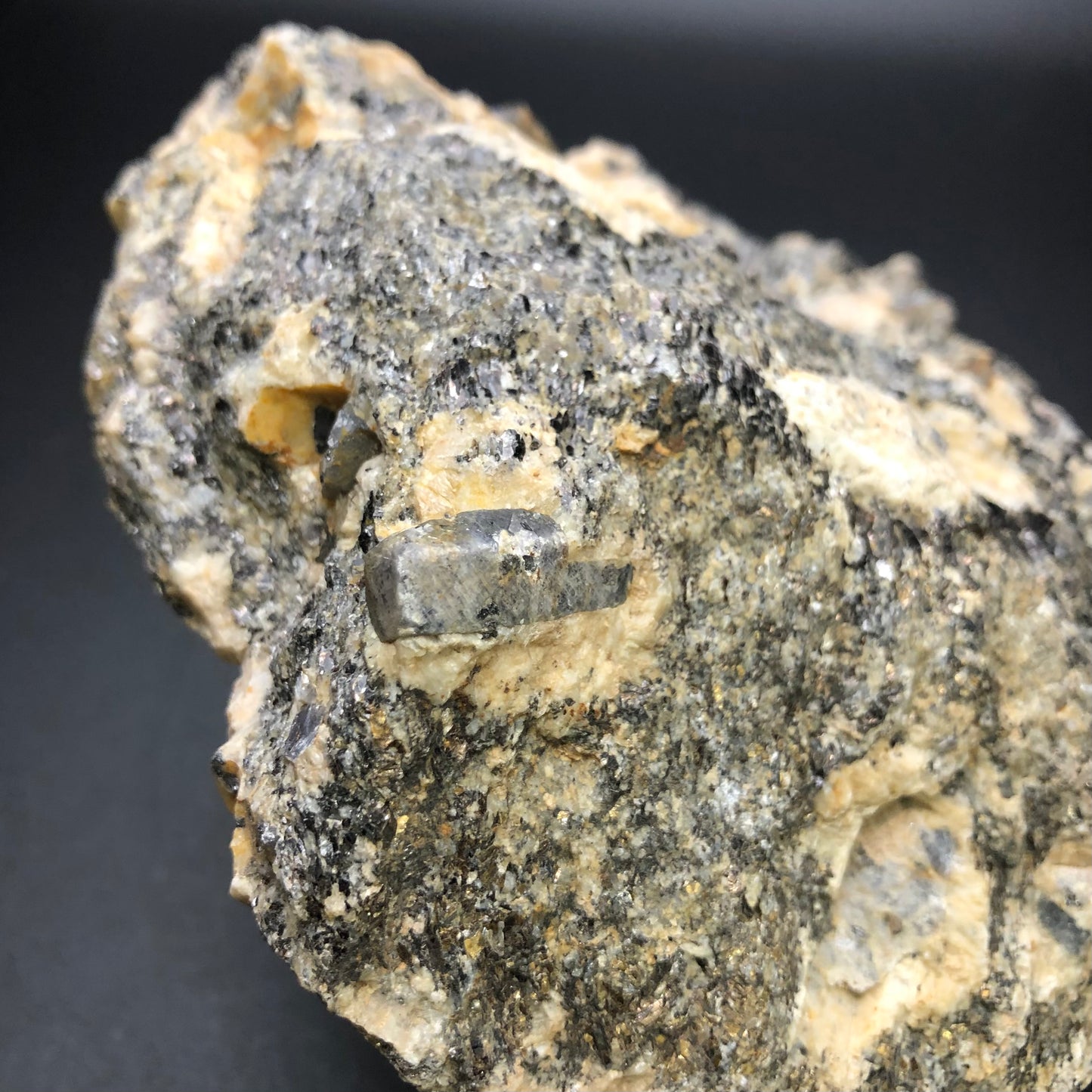 A detailed shot of a rugged piece from The Crystalary's AUCTION collection, featuring corundum from the Elk Creek Corundum Deposit in Gallatin County, Montana. It highlights gray, black, and white mineral patterns with a textured surface, where corundum crystals stand out against a dark backdrop.