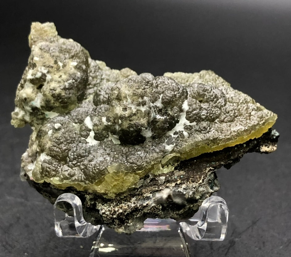 A textured, irregularly shaped rock with a rough, bubbly surface is displayed on a clear plastic stand against a dark background. This Cuprian Smithsonite, featured by The Crystalary from Arizona's well-known 79 Mine in the Chilito region of Gila County, is predominantly gray with subtle hints of pale yellow and green.