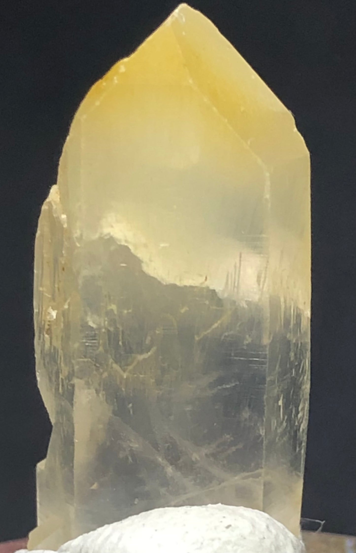 A translucent Mango Quartz crystal from The Crystalary, titled AUCTION- Quartz w/ Halloysite- Cabiche, Quípama, Western Boyacá Province, Colombia, features a pointed tip and visible internal striations. It is set against a dark background and rests on a white mineral base.