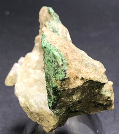 A close-up of a rough mineral specimen resembling Uvarovite Garnet in beige and green hues showcases its earthy textures against a dark background. This druzy stone, from The Crystalary, is possibly sourced from the rich geology of Magog, Quebec, Canada.