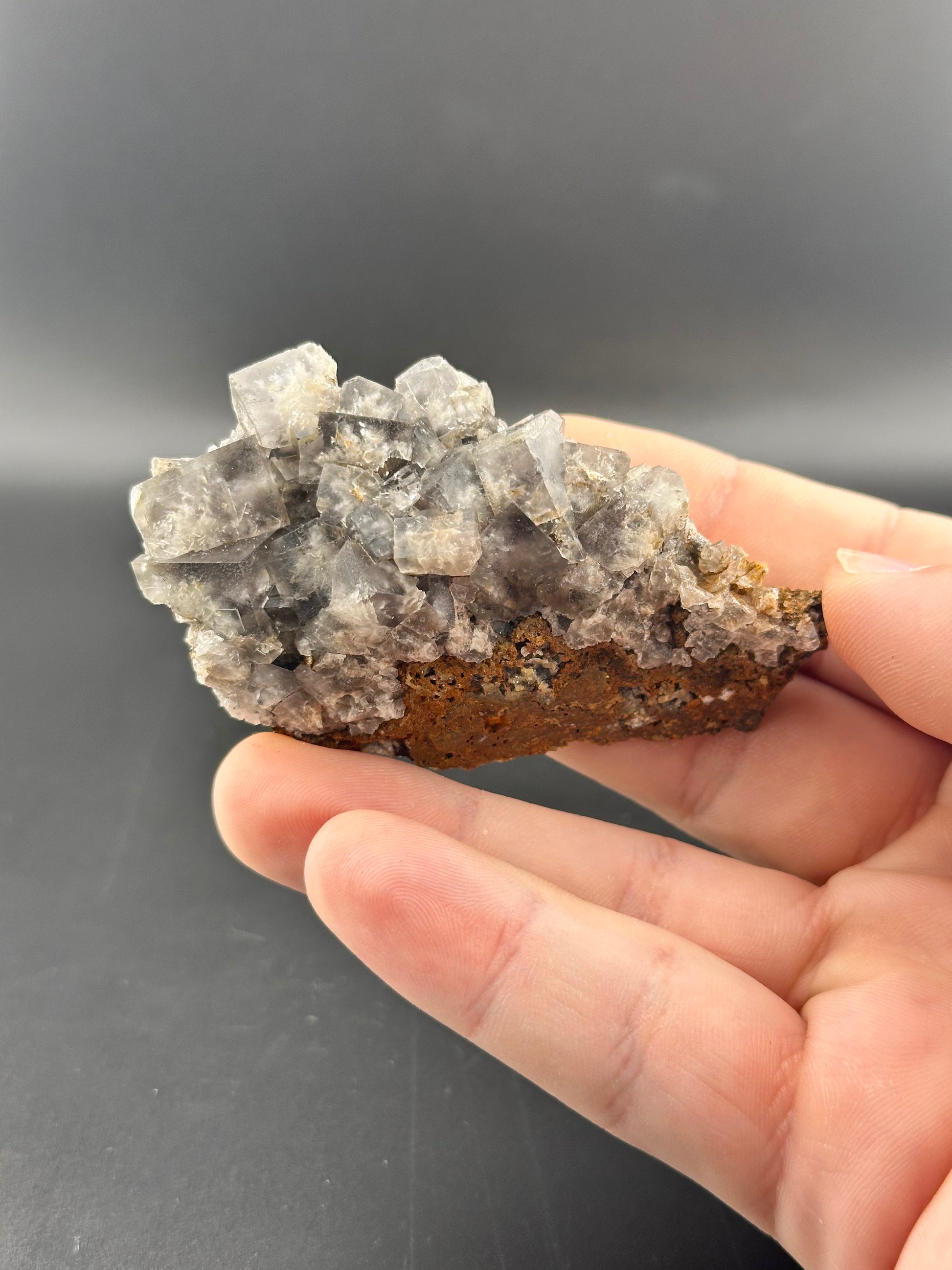 Against a dark backdrop, a hand showcases a gray crystal geode by The Crystalary, reminiscent of the LIVE- Karen- 12/14/24, with its angular formations and brown base.