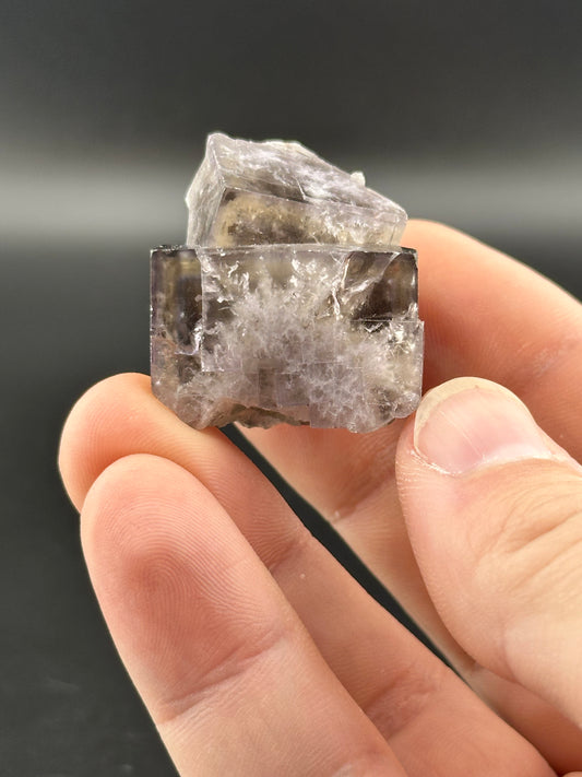 A hand gently holds The Crystalary's $65 LIVE- Mandyfitz- 12/14/24 mineral crystal from the Cromwell Pocket, highlighting its raw, translucent cubic form. Its smoky, grayish-clear hues contrast beautifully against a simple dark background.