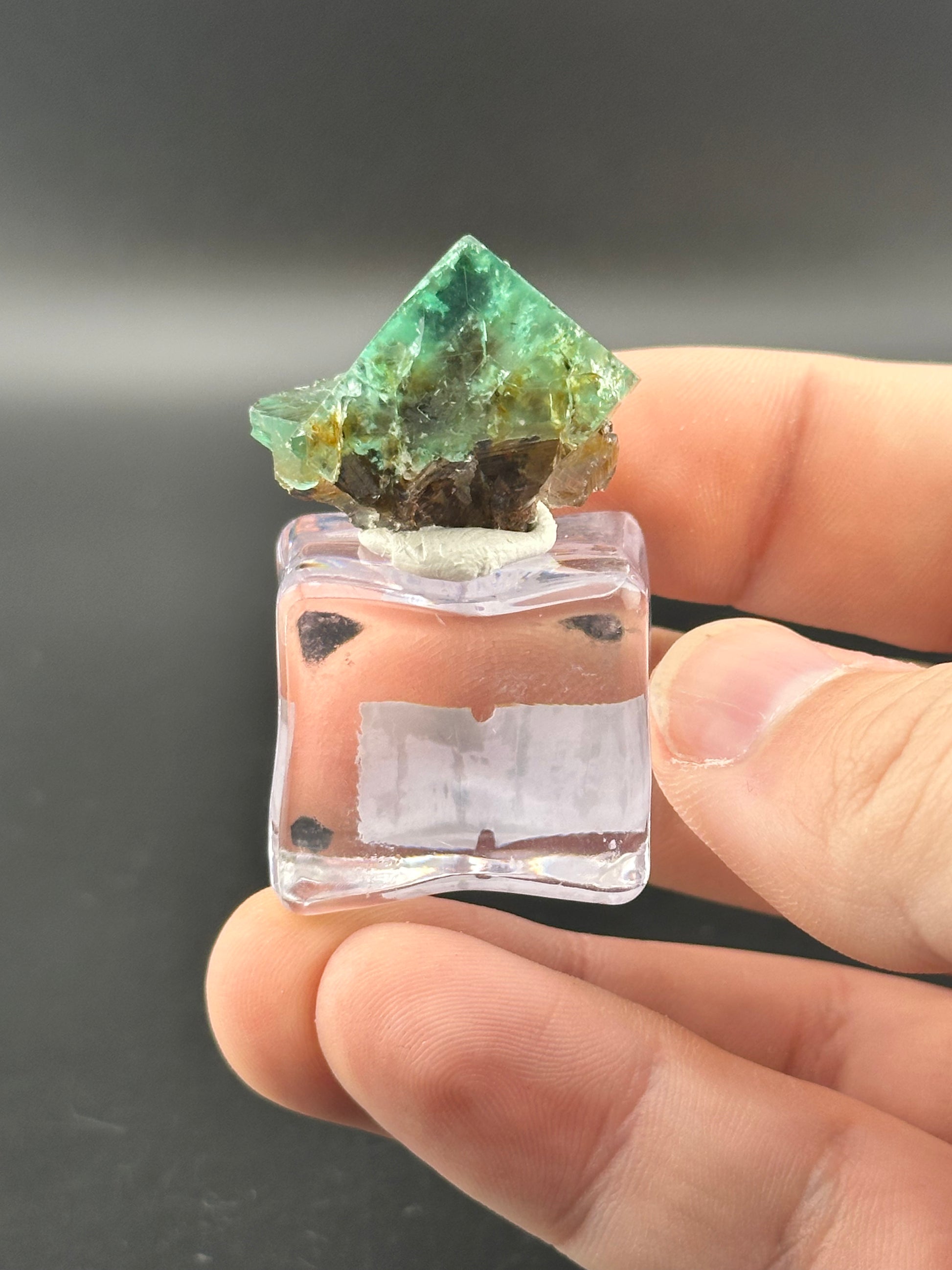 A hand holds a small mineral specimen, the green pyramid-shaped crystal atop it is from The Crystalary's LIVE- Chaos_Supervisor- 12/14/24, set on a clear base. The dark backdrop highlights its intricate structure, evoking the serene mystery of a Hidden Forest.