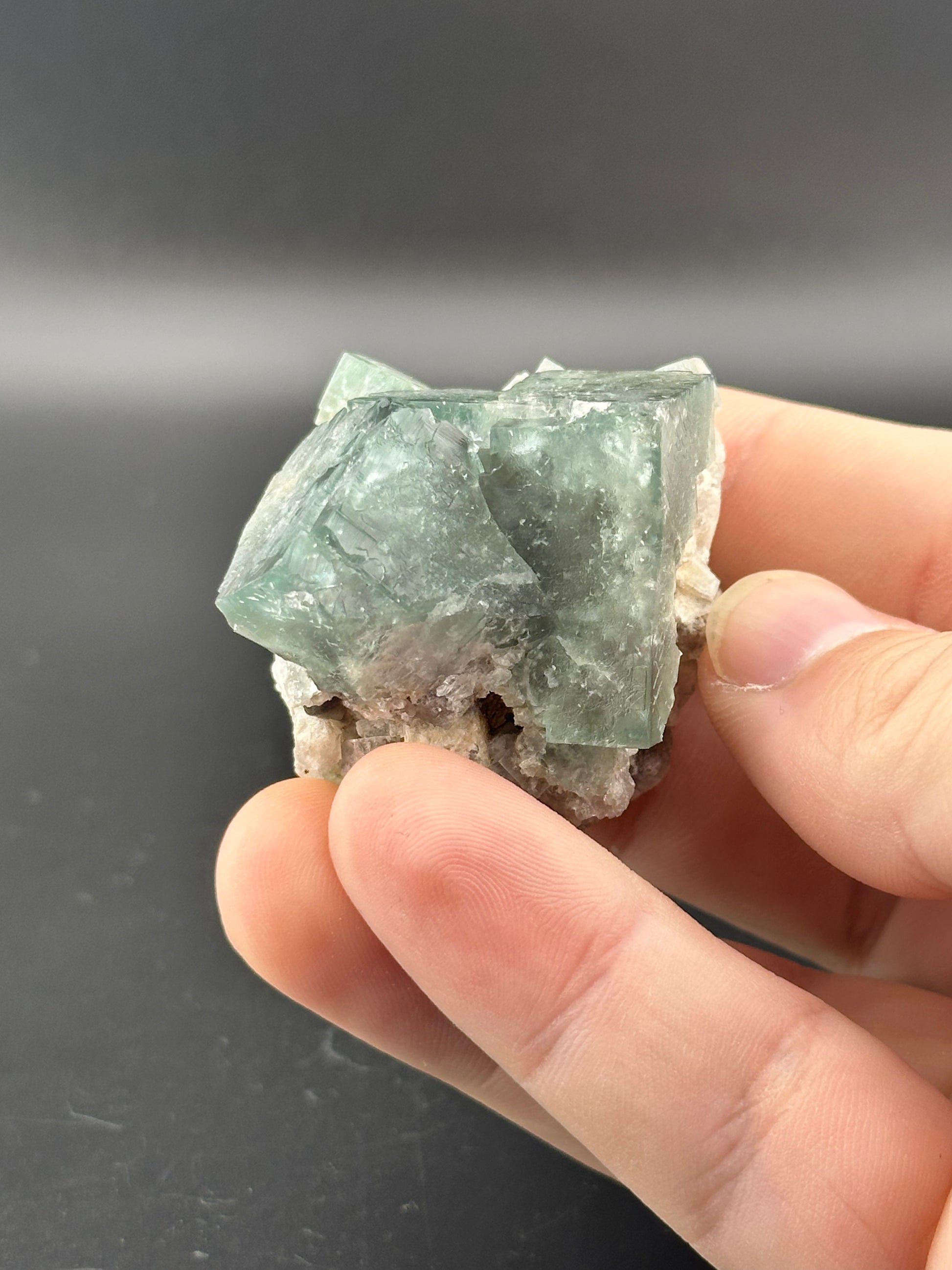 A person holds the LIVE- Chaos_Supervisor- 12/14/24 from The Crystalary, a greenish crystal with a rough, translucent surface against a dark background. The hand's fingers encircle it gently.